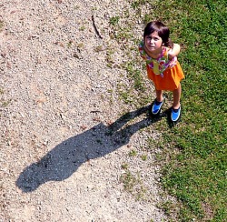 A young girl looks up at the camera, her shadow displayed prominently on the ground in front of her